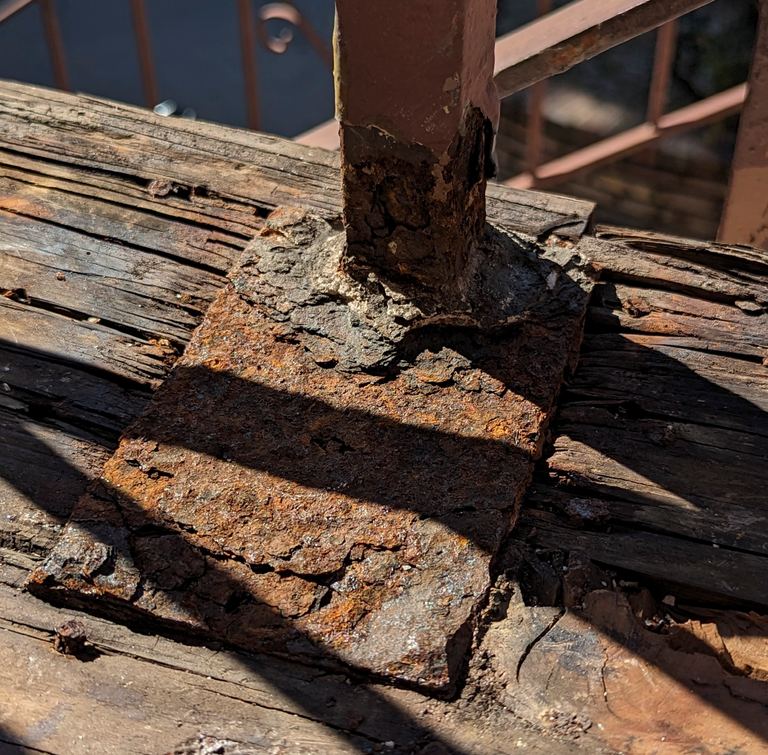 Deteriorated railing post welded to a rusted base plate on a rotted wood ledger
